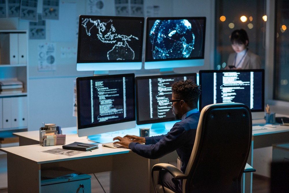 Cybersecurity specialist sits at a desk with five screens monitoring threat levels. 