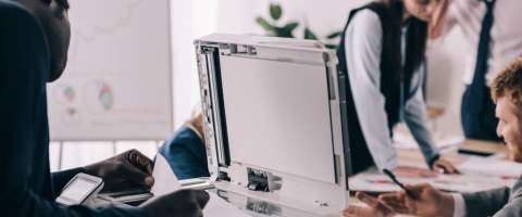 Business team collaborating in a modern workspace with an office copier in use; one person is making copies while others discuss charts and data at a table with a whiteboard in the background.