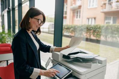 Happy female employee raises the lid on a multifunction printer to make copies symbolizing the importance of copier maintenance.
